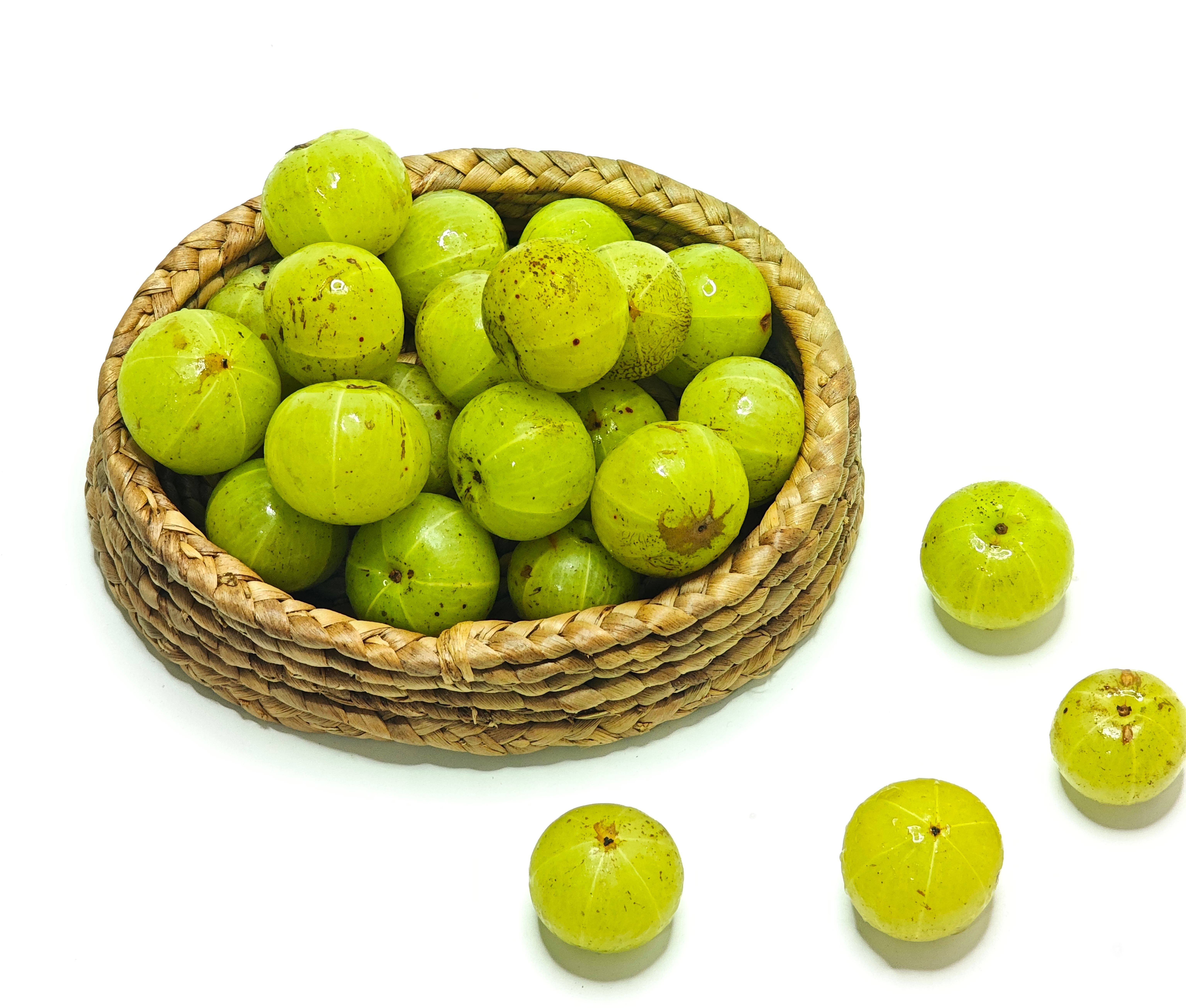Fresh pale green amla berries (Indian gooseberries) with faint vertical ridges and natural surface markings, piled in a light brown woven basket on a clean white background, with several loose fruits scattered nearby. 